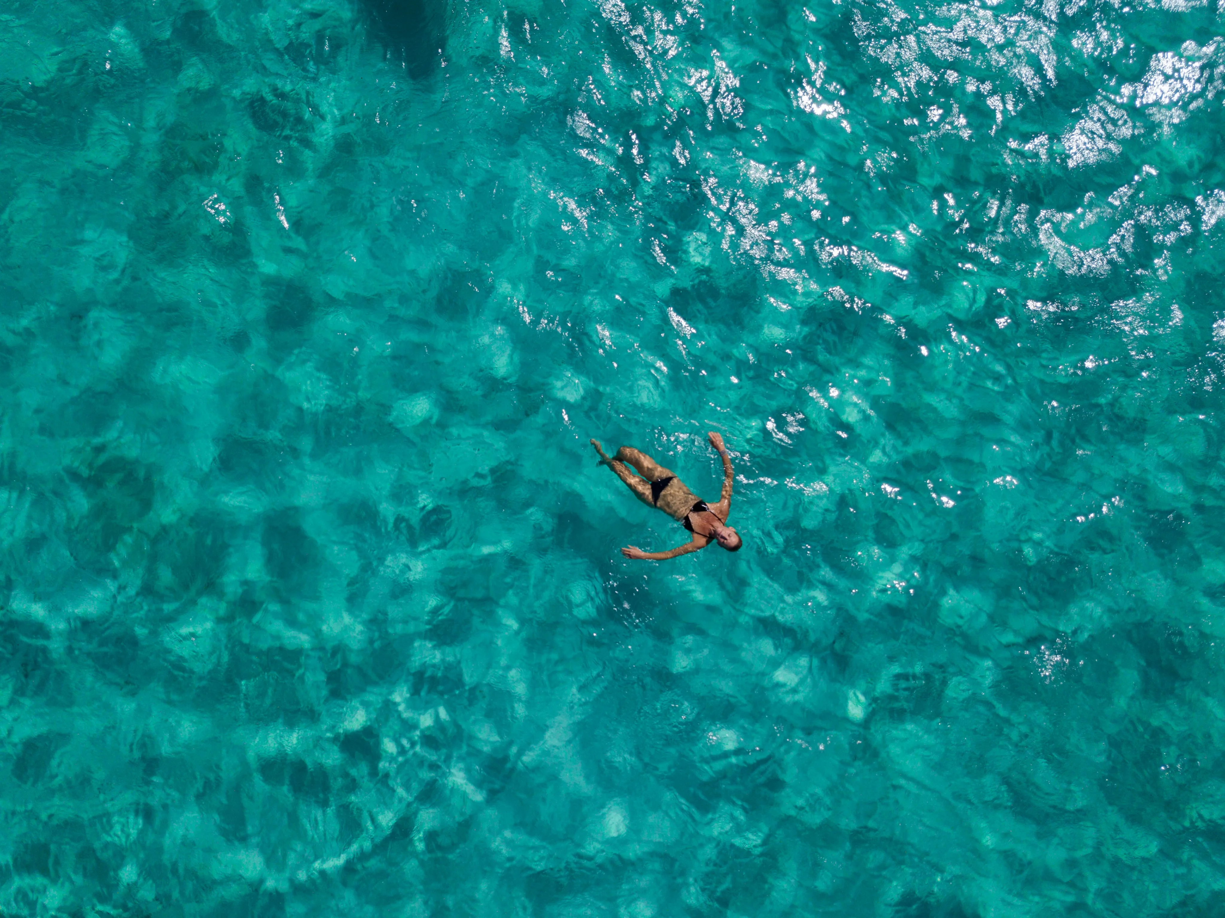 A person floats on their back in clear, turquoise ocean water. The water's surface glistens under the sunlight, creating a serene, tropical scene. The person is relaxed, arms outstretched, and appears to be enjoying the calmness of the sea—opting to stay in this moment of pure tranquility.
