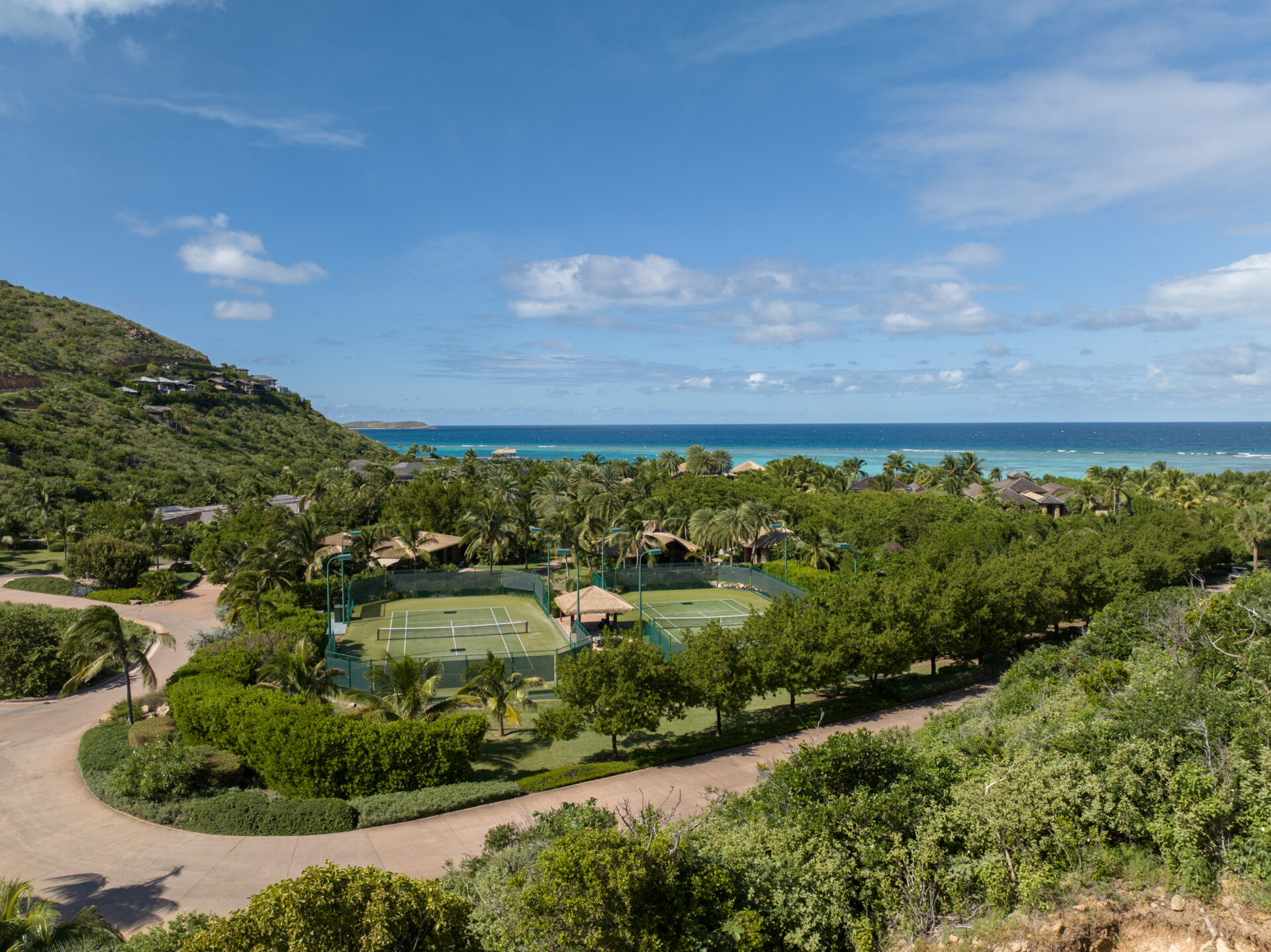 An aerial view of the lush coastal landscape at Atlantic Ridge features multiple tennis courts surrounded by green trees. In the background, the turquoise ocean meets the clear blue sky with a few scattered clouds. Rolling hills and scattered homesites complement this serene setting.