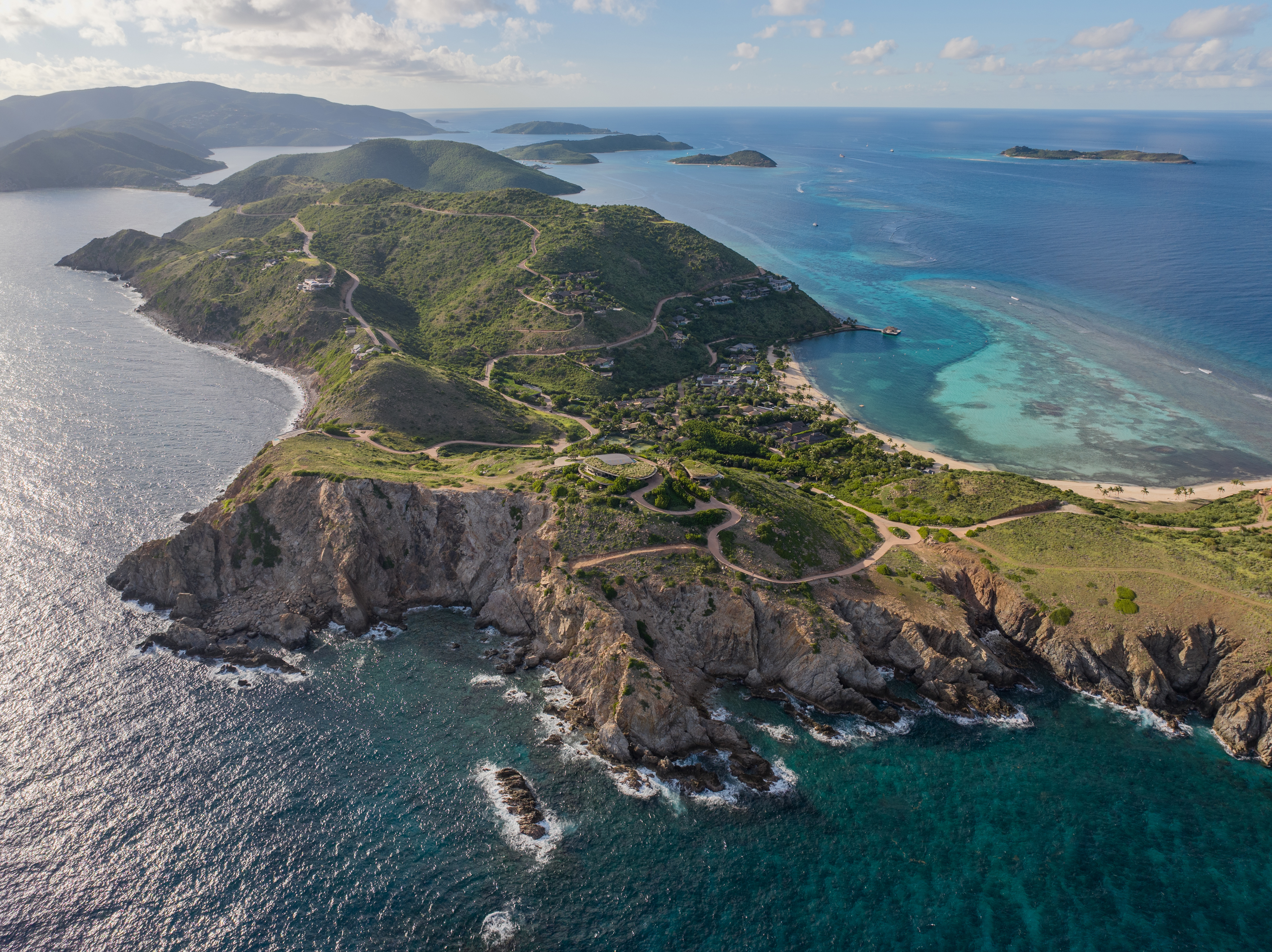 Aerial view of a picturesque island with lush green hills and rugged cliffs surrounded by vibrant blue Atlantic Ridge waters. The island features winding roads, small buildings, and clear shallow reefs near the shoreline. Other small islands are visible in the background.