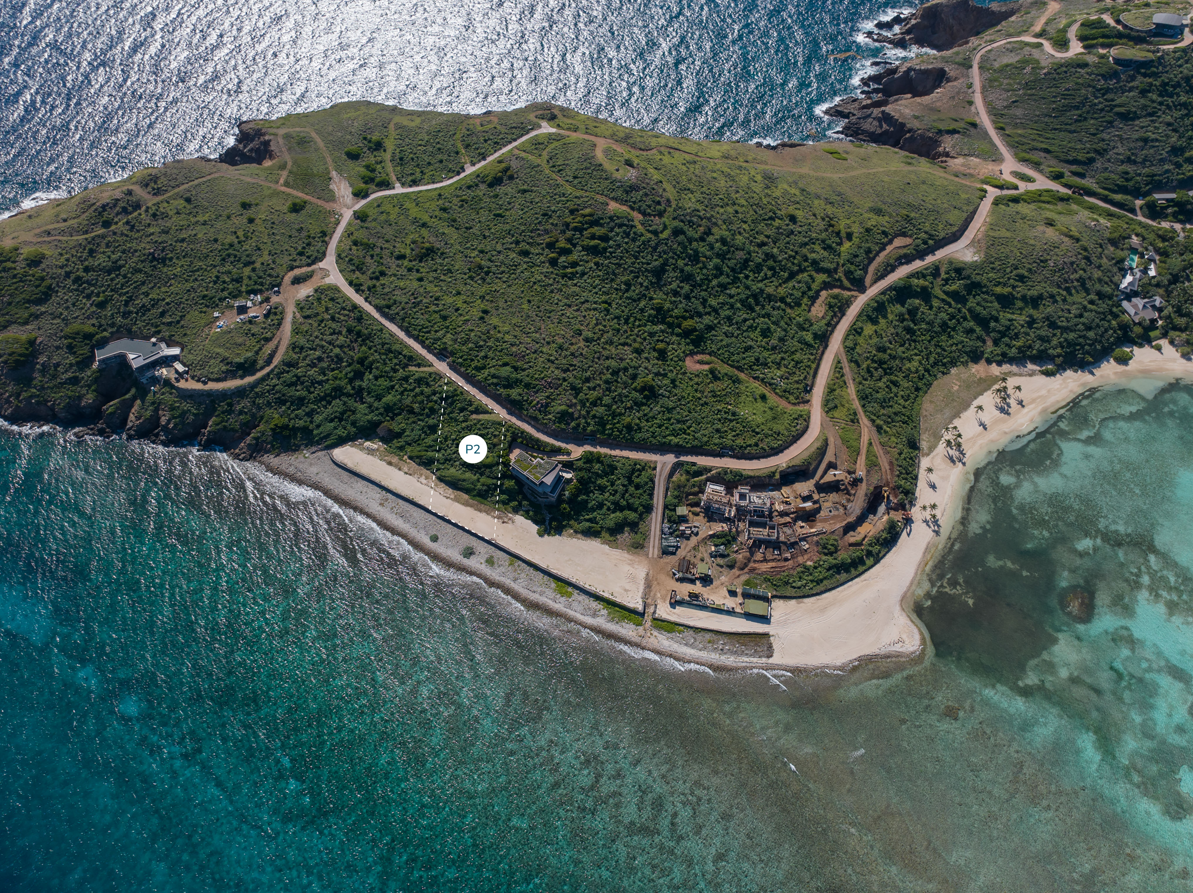 Aerial view of a coastal peninsula featuring a curving road network surrounding a green landscape, sandy beaches, and clear, shallow waters. There are a few scattered homesites and a construction site near the shore. The ocean with gentle waves is in the background.