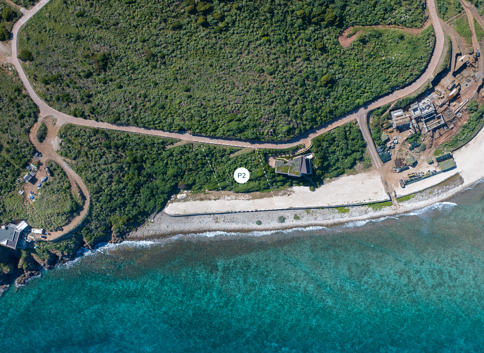 Aerial view of a coastal area featuring clear turquoise waters, a sandy beach, and dense green vegetation. Near the shoreline, next to a small road on the peninsula, there's a homesite marked with a white circle labeled "P2".
