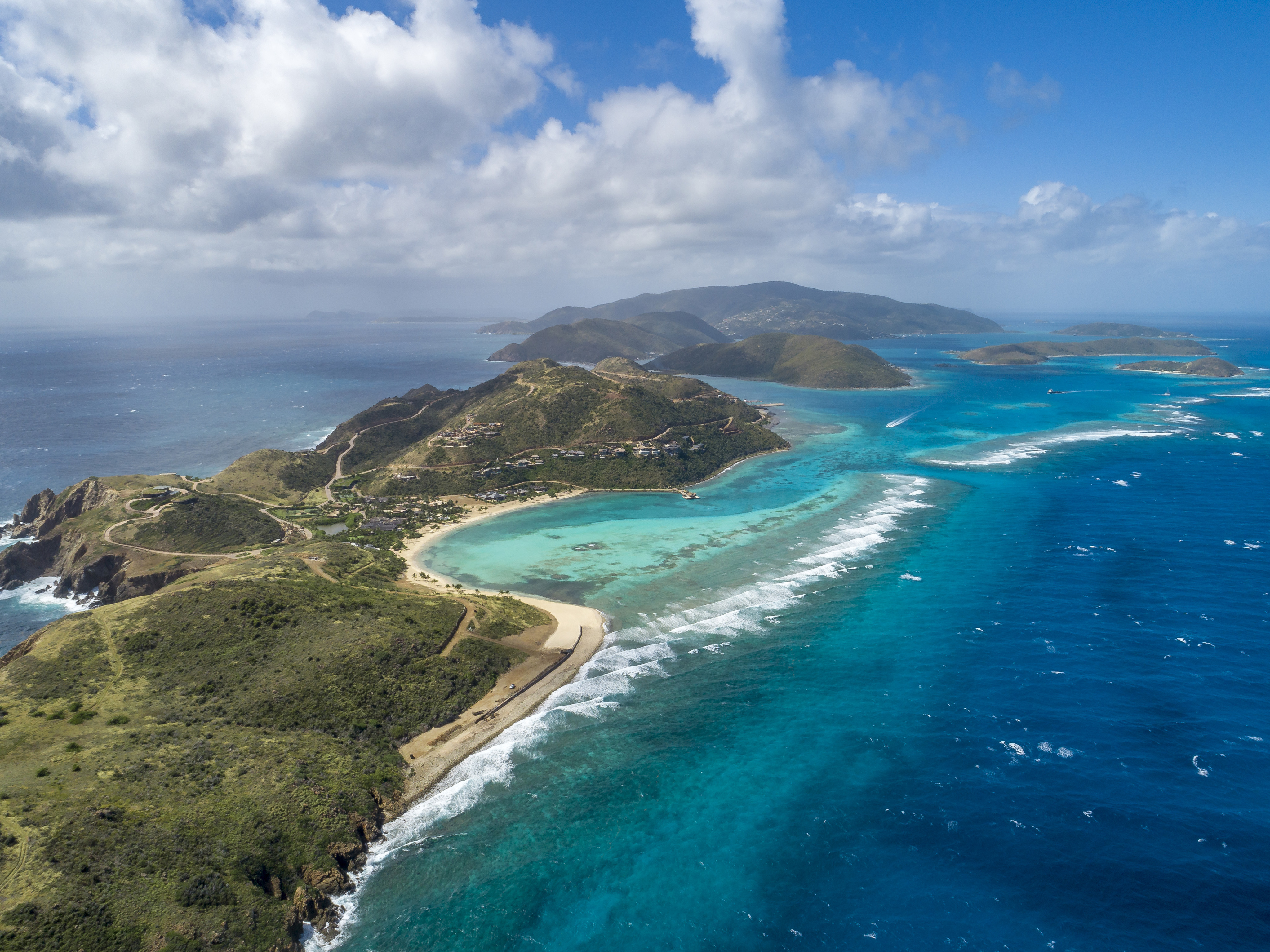Aerial view of a tropical Island Homesite P2 with lush green hills, sandy beaches, and turquoise waters. The shoreline curves gently around the peninsula, bordered by coral reefs and shallow seas. The sky is partly cloudy, and other islands are visible in the distance across the ocean.