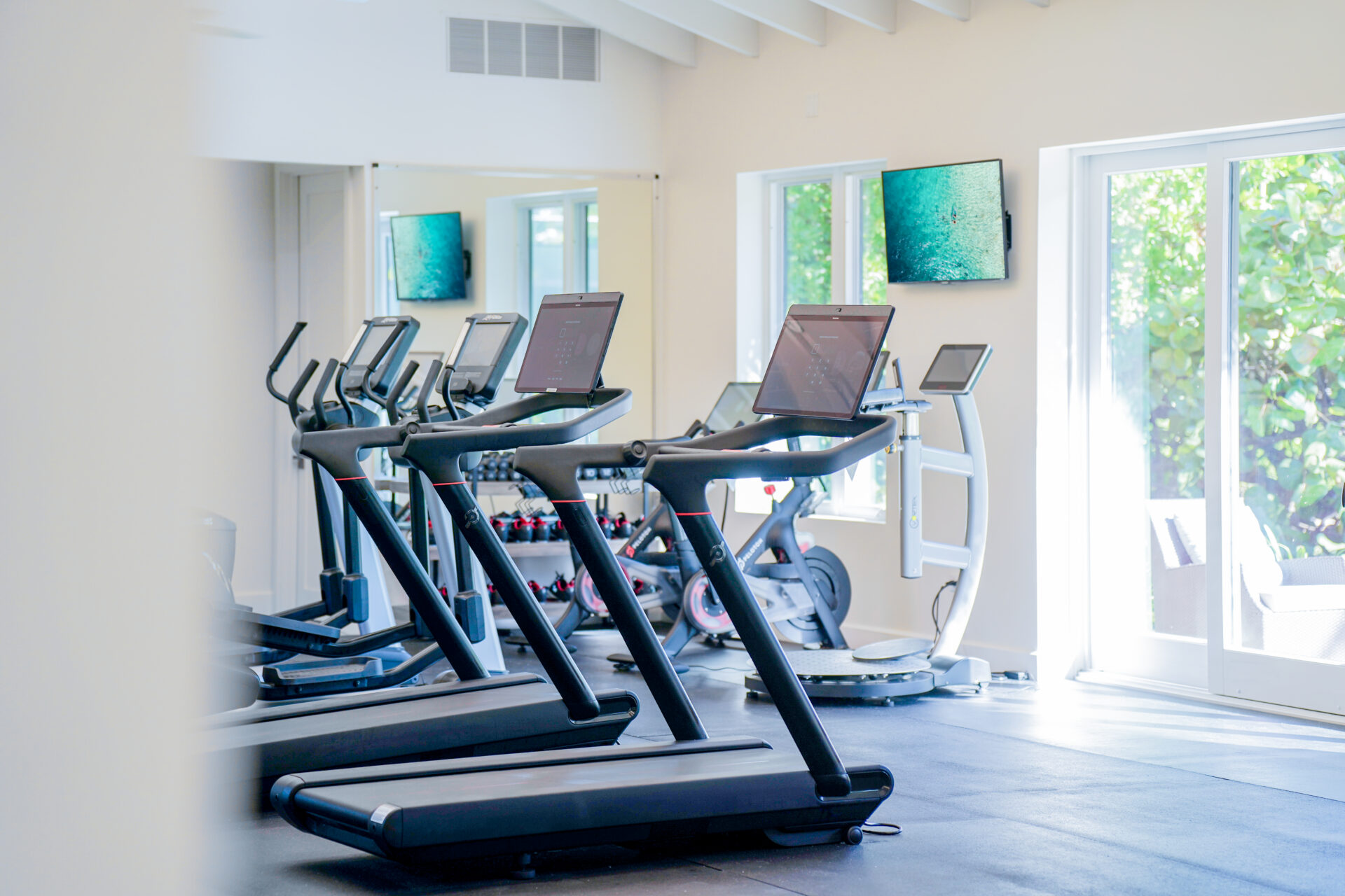A well-lit health studio with several treadmills facing a wall-mounted TV and mirrors. There are large windows on the right letting in natural light, promoting wellness. A stationary bike and other equipment are in the background.