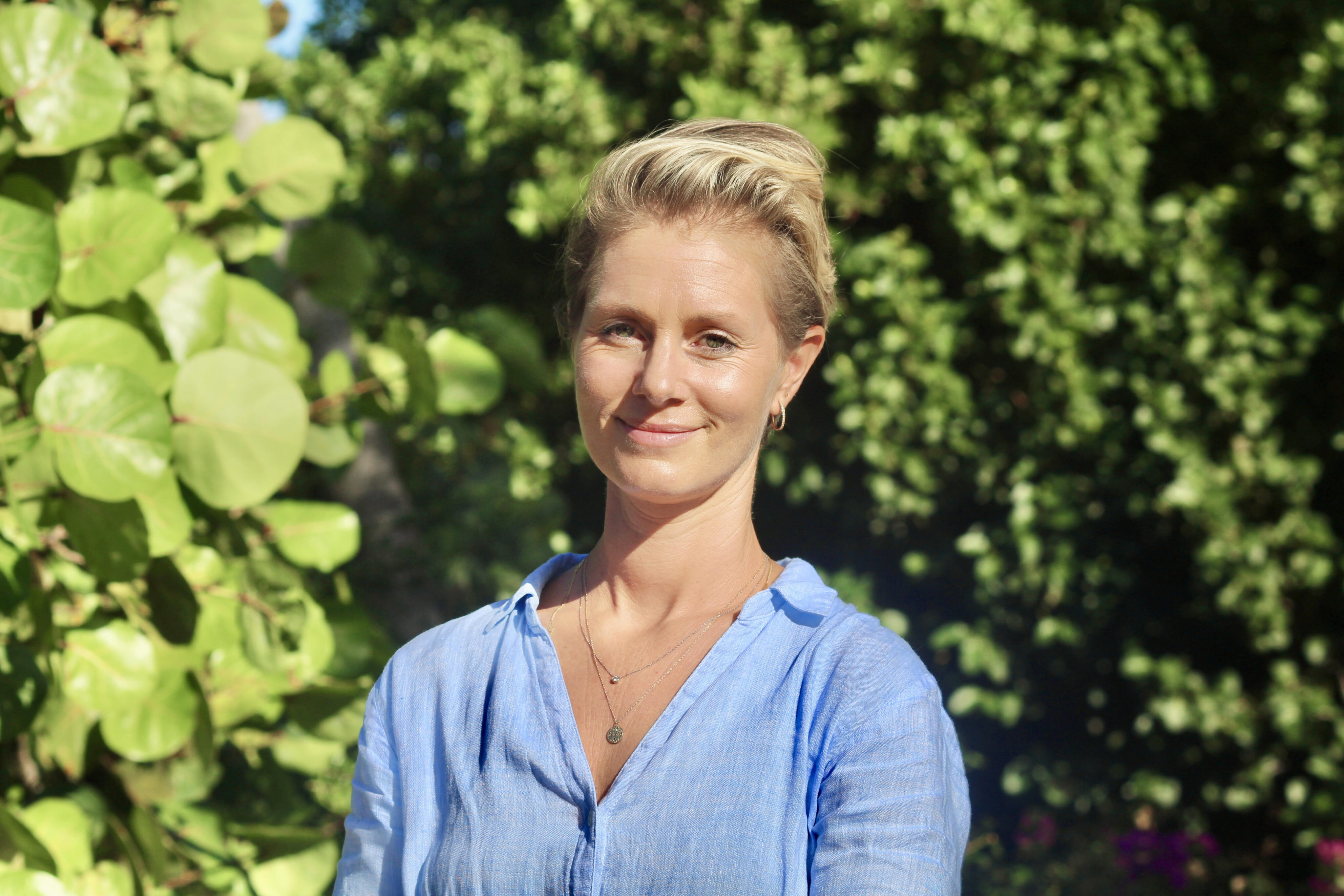 Emily Oakes, a woman with short blonde hair, stands outdoors in front of green foliage. She wears a light blue blouse and smiles gently at the camera. The bright lighting suggests a sunny day. Emily adorns two delicate necklaces and small hoop earrings.