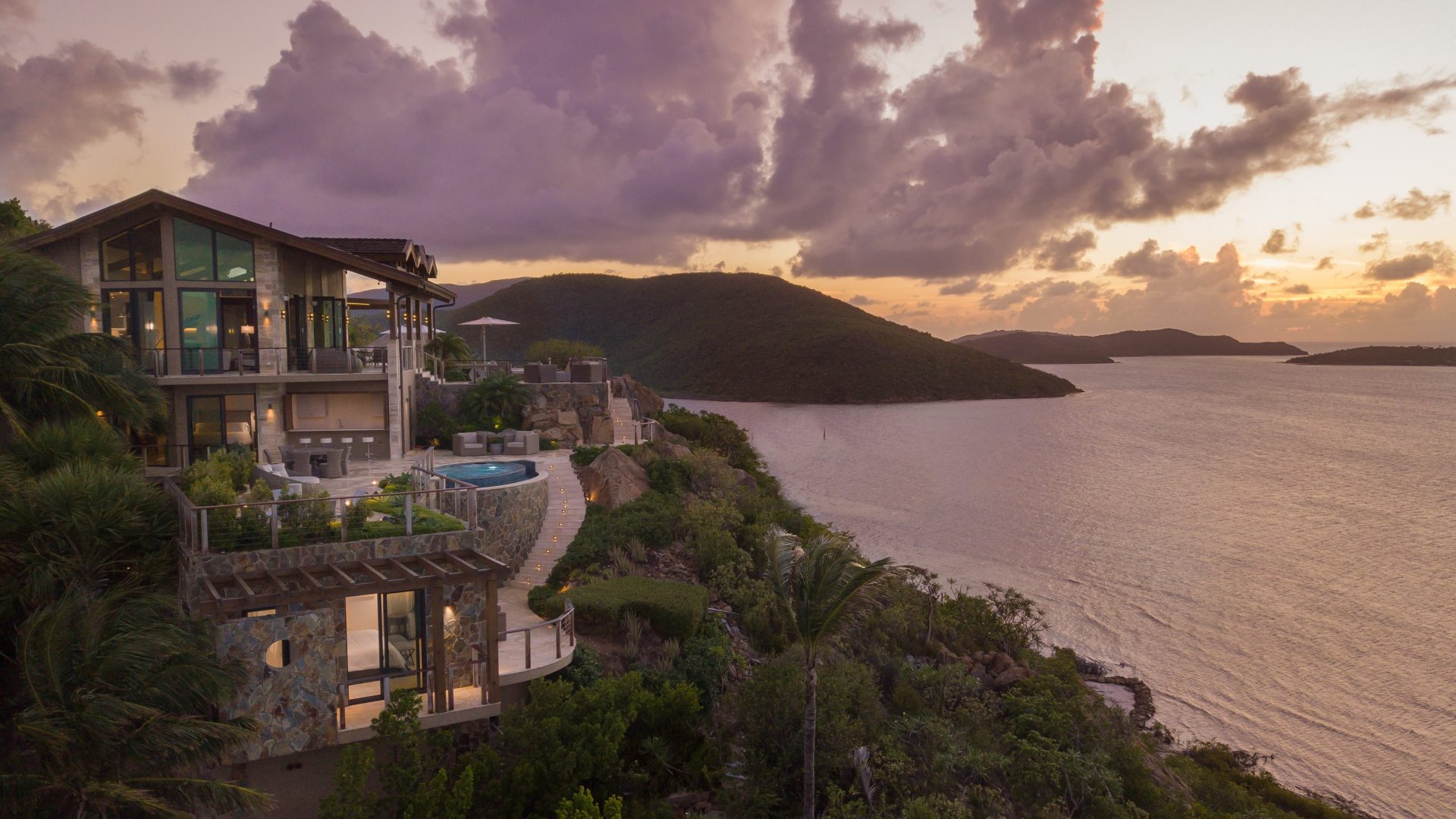 A luxurious multi-story house with large windows overlooks a serene body of water at sunset. The house features a pool and outdoor seating area on the cliffside, surrounded by lush greenery. The sky is filled with dramatic clouds and the scene is tranquil and picturesque—villas and homesites for sale in the BVI.
