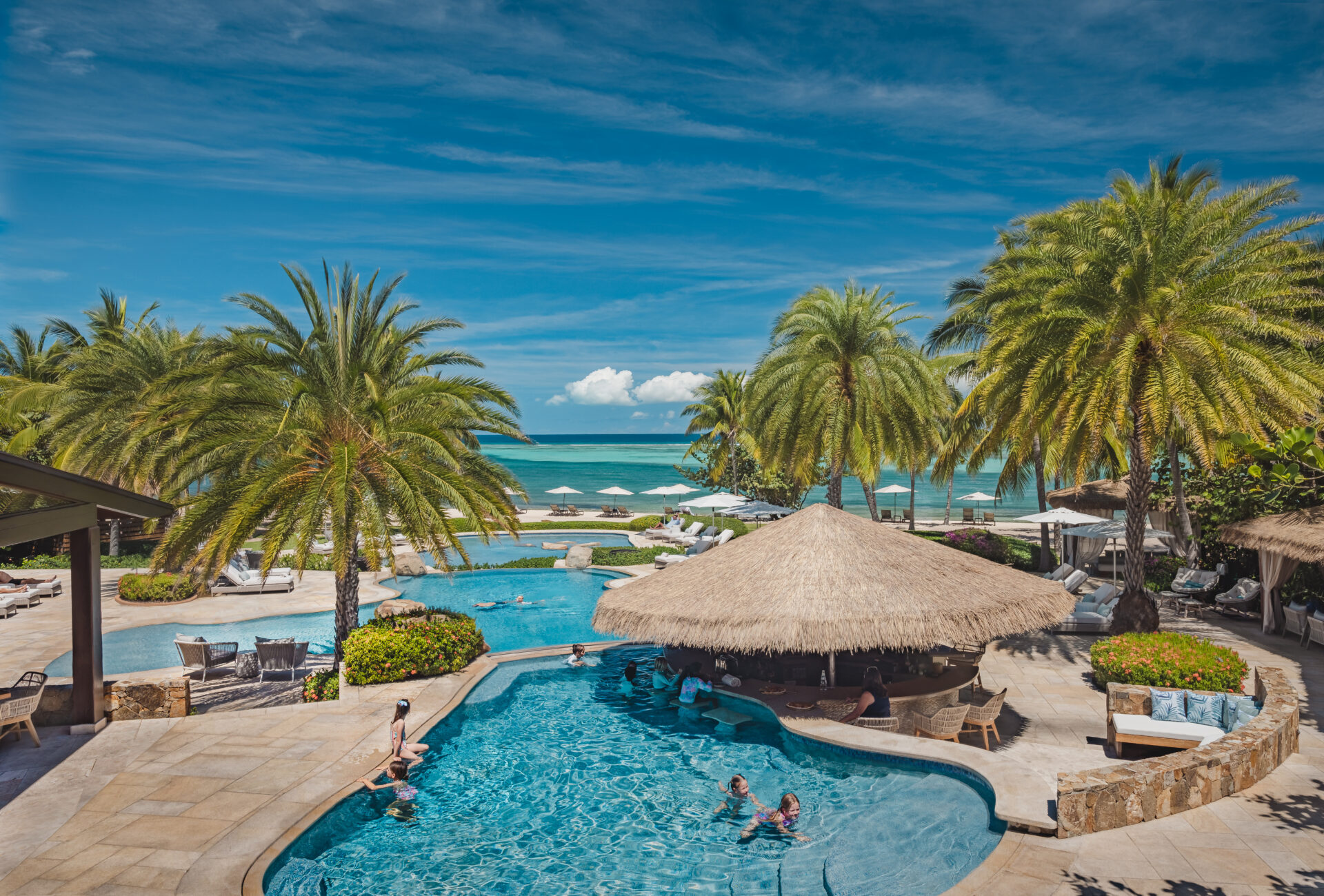 A luxurious tropical resort, Oil Nut Bay, features multiple swimming pools surrounded by lush palm trees. Guests relax in and around the pools, including a swim-up bar with thatched roofing. The resort overlooks a turquoise ocean under a clear blue sky.