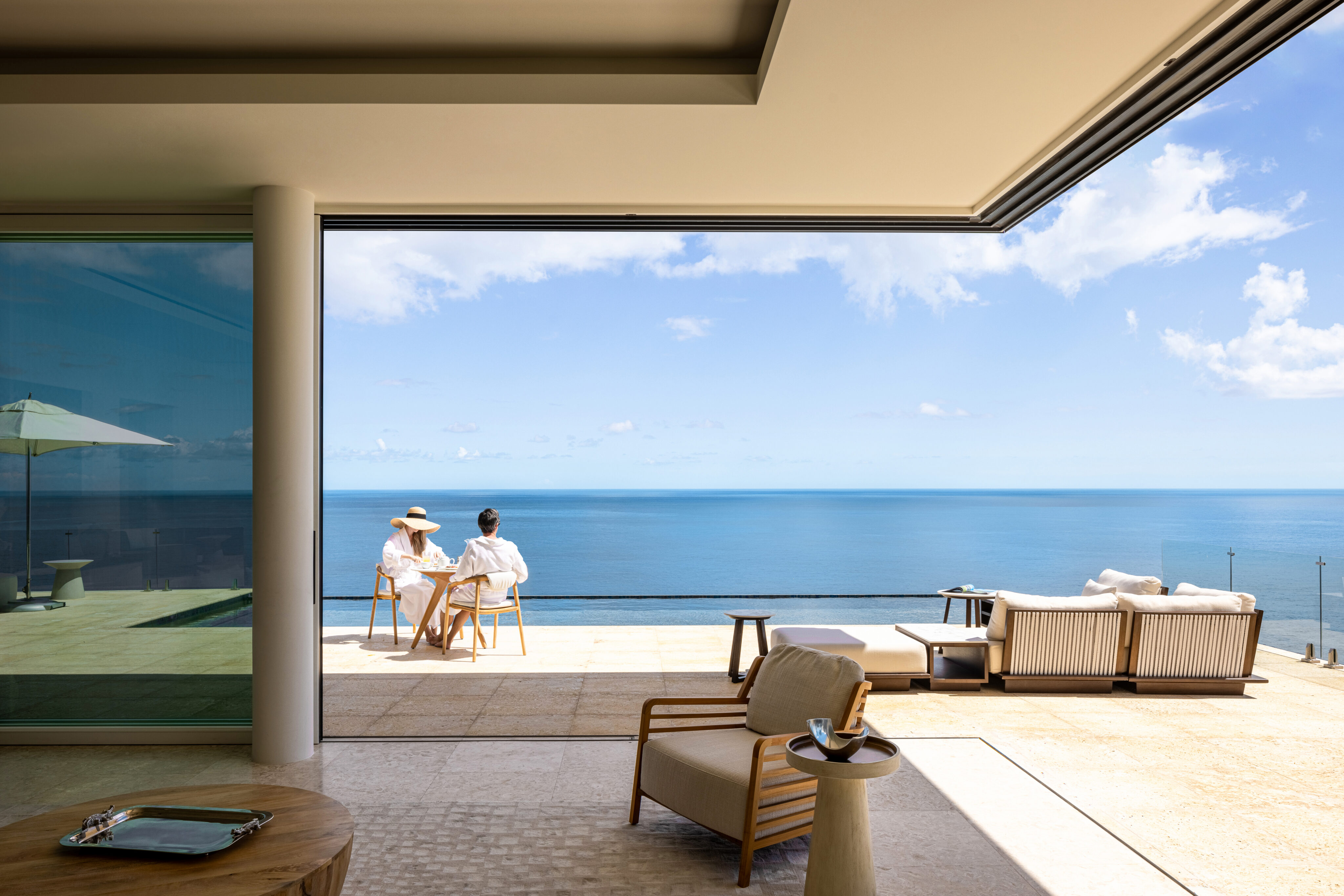 Two people in white robes sit at a table on a spacious patio with a sea view in Oil Nut Bay. Modern furniture, including chairs and a sofa, is placed around the area. A large glass window is on the left, and the clear blue sky meets the ocean horizon in the background.