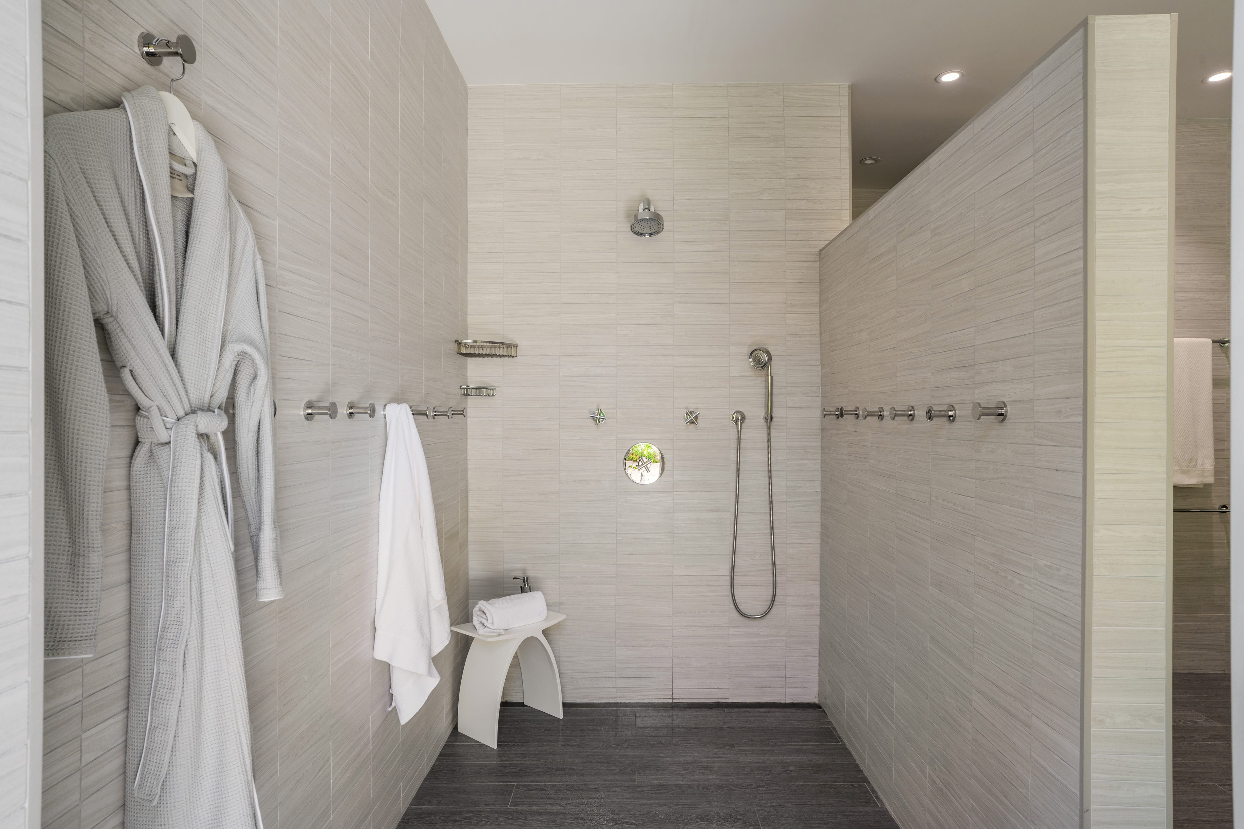 A spacious modern shower room at Nonamé Villa features light-colored textured walls, a wall-mounted showerhead, handheld shower, robe hooks with a white robe and towel hanging, and a small stool with rolled towels. The floor is covered with dark tiles.