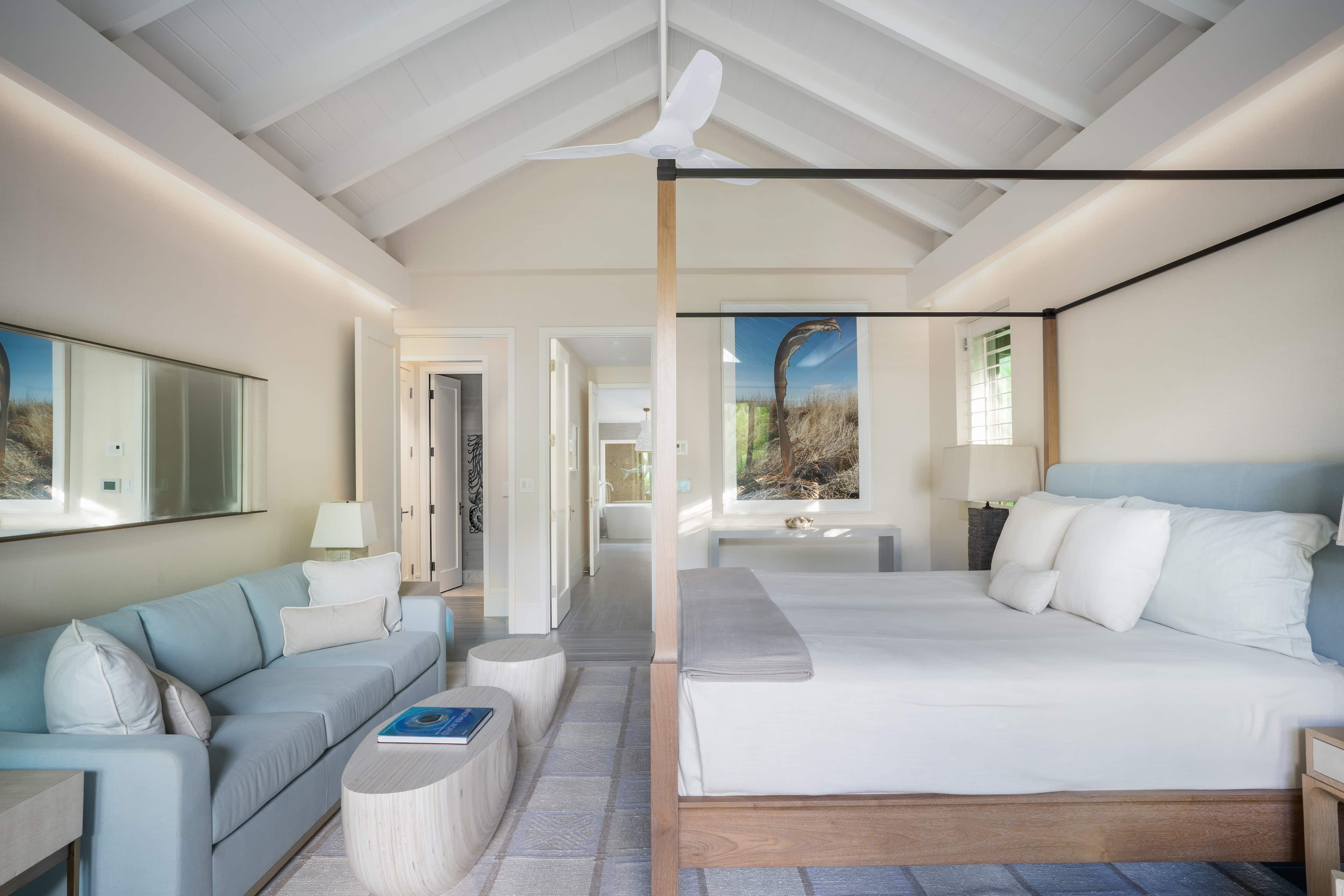 A spacious, light-filled bedroom at Nonamé Villa with a vaulted ceiling features a large bed with plush pillows and a white canopy frame, a cozy sitting area with a blue sofa and wooden coffee table, a wall-mounted TV, and serene coastal artwork. Neutral tones dominate the decor.