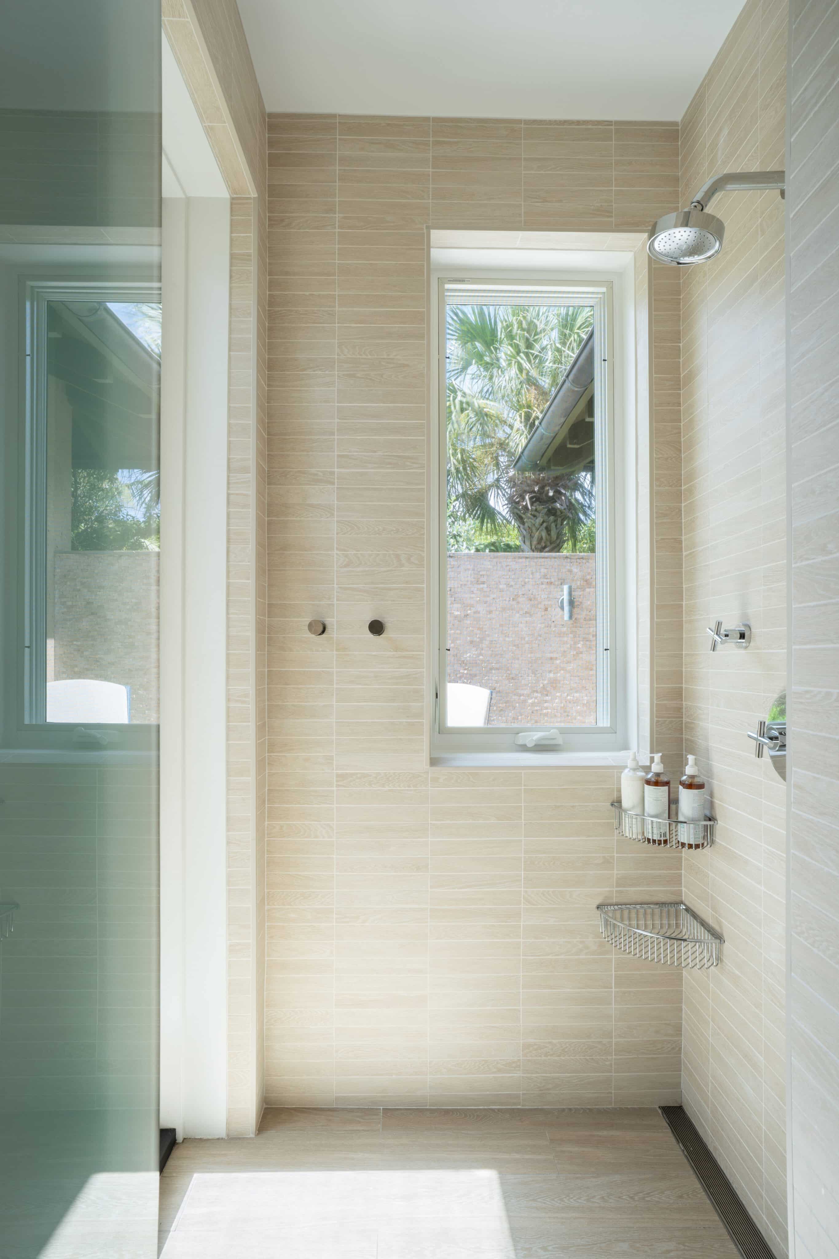 A modern, beige-tiled bathroom in Nonamé Villa with a large window. The shower area features a chrome showerhead and a metal corner shelf holding bottles. A partial glass partition separates the shower from the rest of the bathroom. Palm trees are visible outside the window.