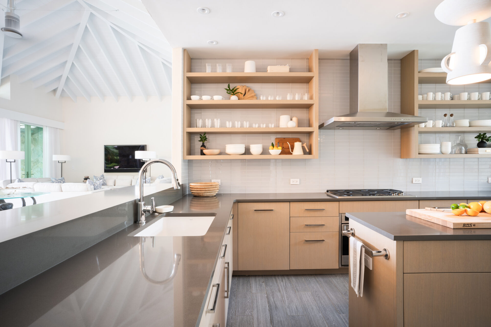 Modern kitchen in Nonamé Villa with light wood cabinets, open shelving displaying white dishes, and stainless steel appliances. A large island features a sink and faucet with a gray countertop. The room is well-lit with natural light from windows and pendant lights above.