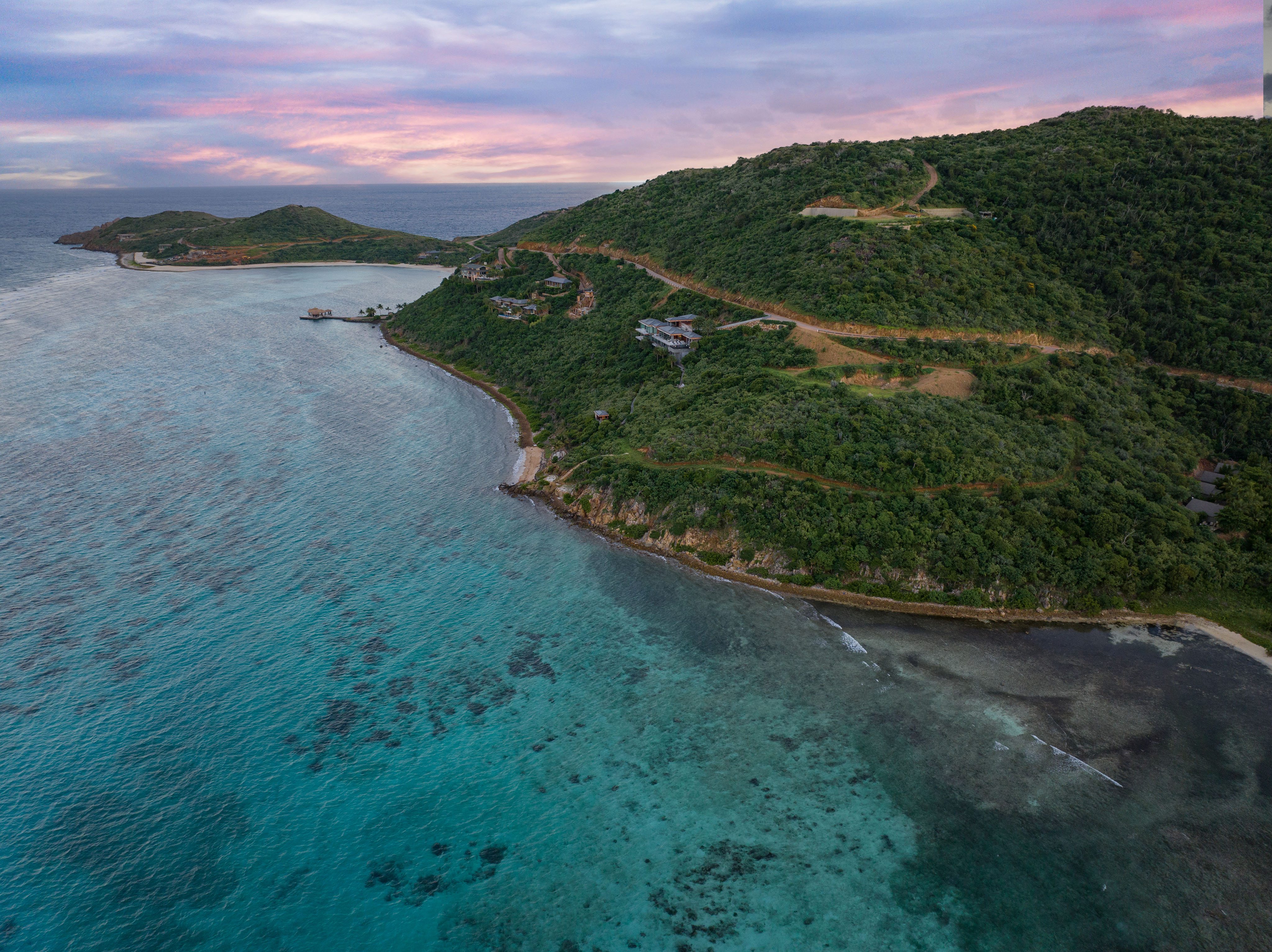 Aerial view of a coastal landscape with lush green hills and clear turquoise waters. The E15 road elegantly winds along the coastline. The sky, a mix of pink and blue hues, suggests either sunrise or sunset. Estate homesites dot the area, and small islands are visible in the distance.