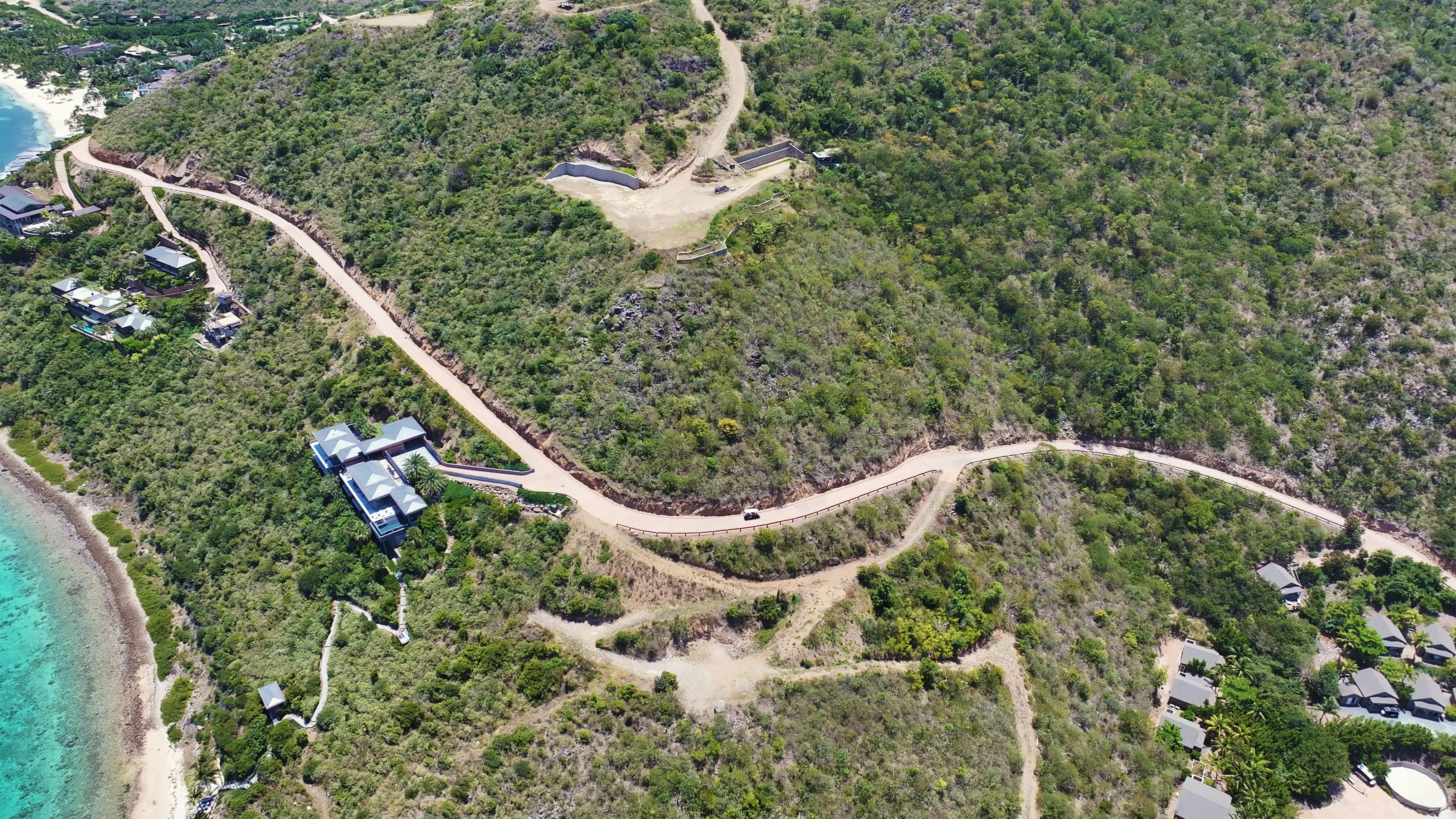 Aerial view of a hilly landscape with a winding road. A modern estate is visible near the road, surrounded by greenery. The coastline with blue water is seen on the left, and several small homesites are scattered across the area.