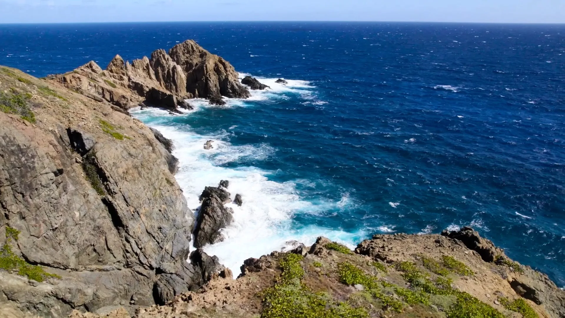 A rocky coastline with jagged cliffs and crashing ocean waves under a clear blue sky, reminiscent of The Point. The deep blue sea churns white foam against the rocks, while sparse greenery clings to the cliff edges near this picturesque homesite.