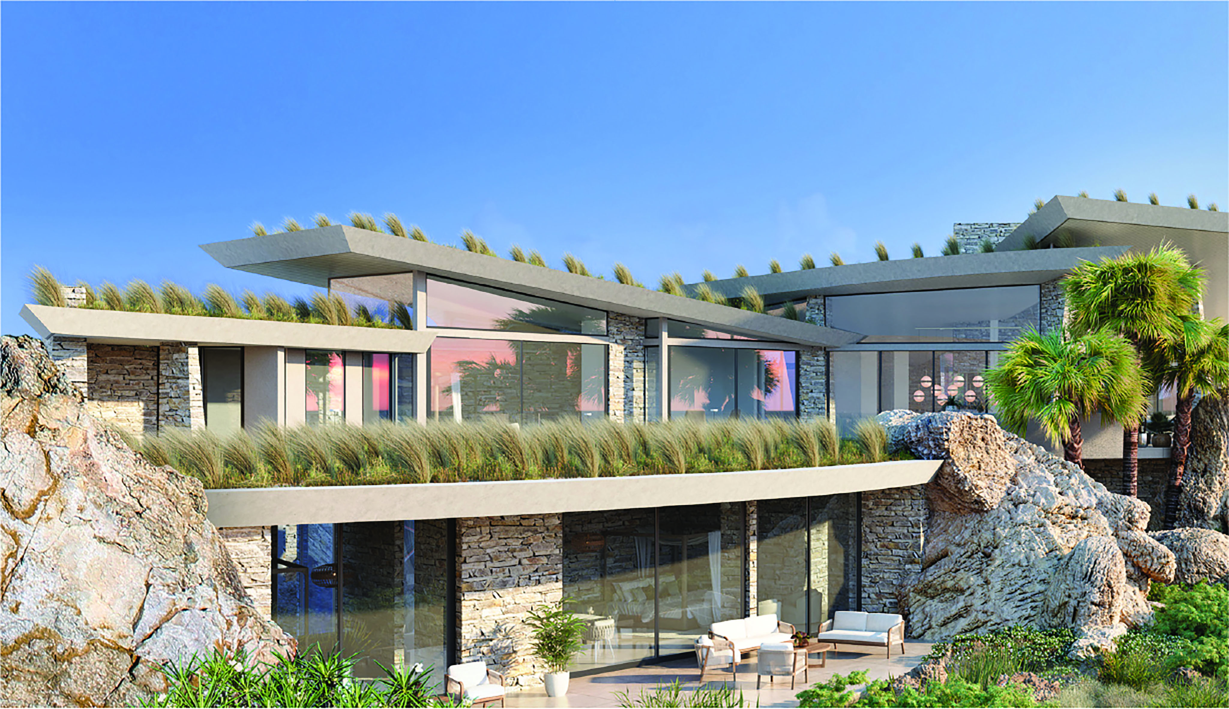 A modern luxury house with large glass windows is nestled on a Boulder Homesite among rocky terrain. The architecture features multiple flat roofs with grass growing on them, blending seamlessly with the natural surroundings. Palm trees are visible nearby under a blue sky.