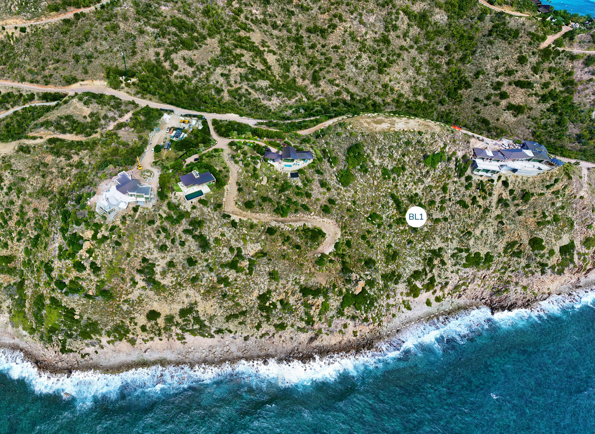 Aerial view of a coastal landscape featuring several boulder homesites nestled among green vegetation on a cliff overlooking the ocean. A dirt road winds through the area, and waves crash against the rocky shore below.