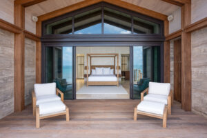 Luxurious Ocean Villa with a modern design, featuring a wooden deck. Two white cushioned chairs face a spacious bedroom with a four-poster bed, large windows providing ample natural light, and an exquisite view of the ocean.
