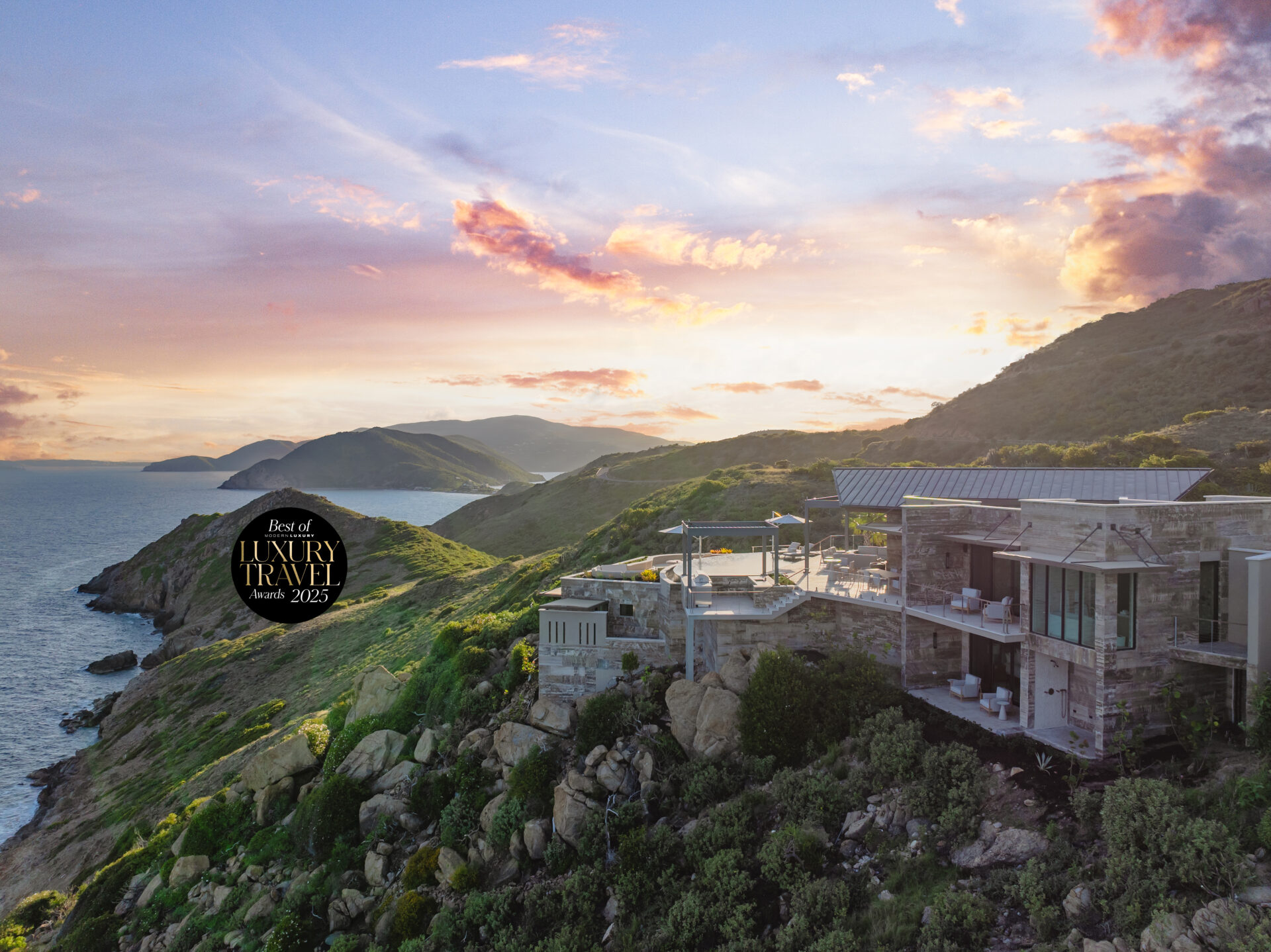 Modern luxury Quintessa Villa perched on a rocky, green hillside overlooking the ocean at sunset, with vibrant clouds in the sky. A "Best of Luxury Travel 2023" emblem is displayed on the left side of the image.