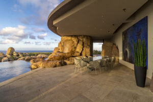 Modern outdoor dining area in Oil Nut Bay featuring a large glass-topped table and white chairs under a curved concrete overhang. This area of the home overlooks a scenic rocky landscape and an infinity pool. A large abstract painting hangs on the wall, with a tall vase of green plants nearby.