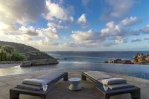 An infinity pool at Oil Nut Bay overlooks a scenic coastal vista in the British Virgin Islands, with two lounge chairs and a small table with drinks on a stone patio. The background features a serene ocean, rocky hills, and a partly cloudy sky at either sunset or sunrise. It’s the perfect home away from home.
