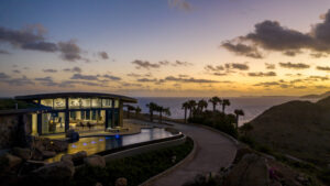 A modern home with large glass windows glows warmly in the evening light, facing a serene ocean view at Oil Nut Bay in the British Virgin Islands. A curved infinity pool mirrors the colors of the setting sun, while palm trees and rugged hills surround the property. The sky is painted with shades of orange and purple.
