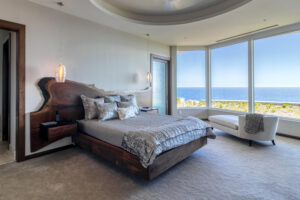 A modern bedroom, reminiscent of a tranquil retreat at Oil Nut Bay in the British Virgin Islands, features a wooden bed with gray bedding and a distinctive wood and resin headboard. Pendant lights illuminate the space, while floor-to-ceiling windows offer a stunning ocean view. A white chaise lounge completes this serene home sanctuary with neutral colors and carpet flooring.