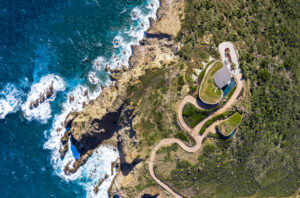 Aerial view of a coastal landscape in the British Virgin Islands featuring a winding pathway leading to a modern building with a green roof. The scene includes rugged cliffs, lush vegetation, and waves crashing against the rocky shoreline of Oil Nut Bay's blue ocean.