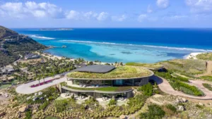 Aerial view of a luxurious, modern home with a green roof overlooking a turquoise coastline in Oil Nut Bay. This luxury escape is surrounded by lush landscaping and hills, offering sweeping views of the ocean and blue sky in the British Virgin Islands.