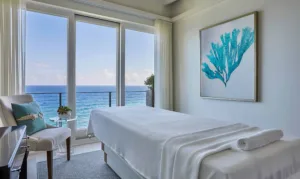 A serene spa room in Oil Nut Bay with a massage table facing floor-to-ceiling windows overlooking the ocean. A rolled towel rests on the table. The room is decorated with turquoise coral artwork and a small side table, perfect for a luxury escape in the British Virgin Islands.