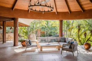 Experience a luxury escape at this outdoor patio, featuring a round wooden roof and modern seating with a sofa, chairs, and coffee table. A circular chandelier adorns the ceiling, surrounded by lush tropical plants. Natural light fills this oasis reminiscent of Oil Nut Bay in the British Virgin Islands.