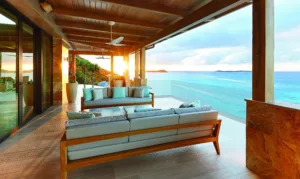 This luxury escape offers a lavish outdoor terrace with wooden furniture and gray cushions, overlooking the vast ocean of Oil Nut Bay in the British Virgin Islands. The sun sets on the horizon, casting a warm glow over the modern space with a wooden ceiling and glass railing.
