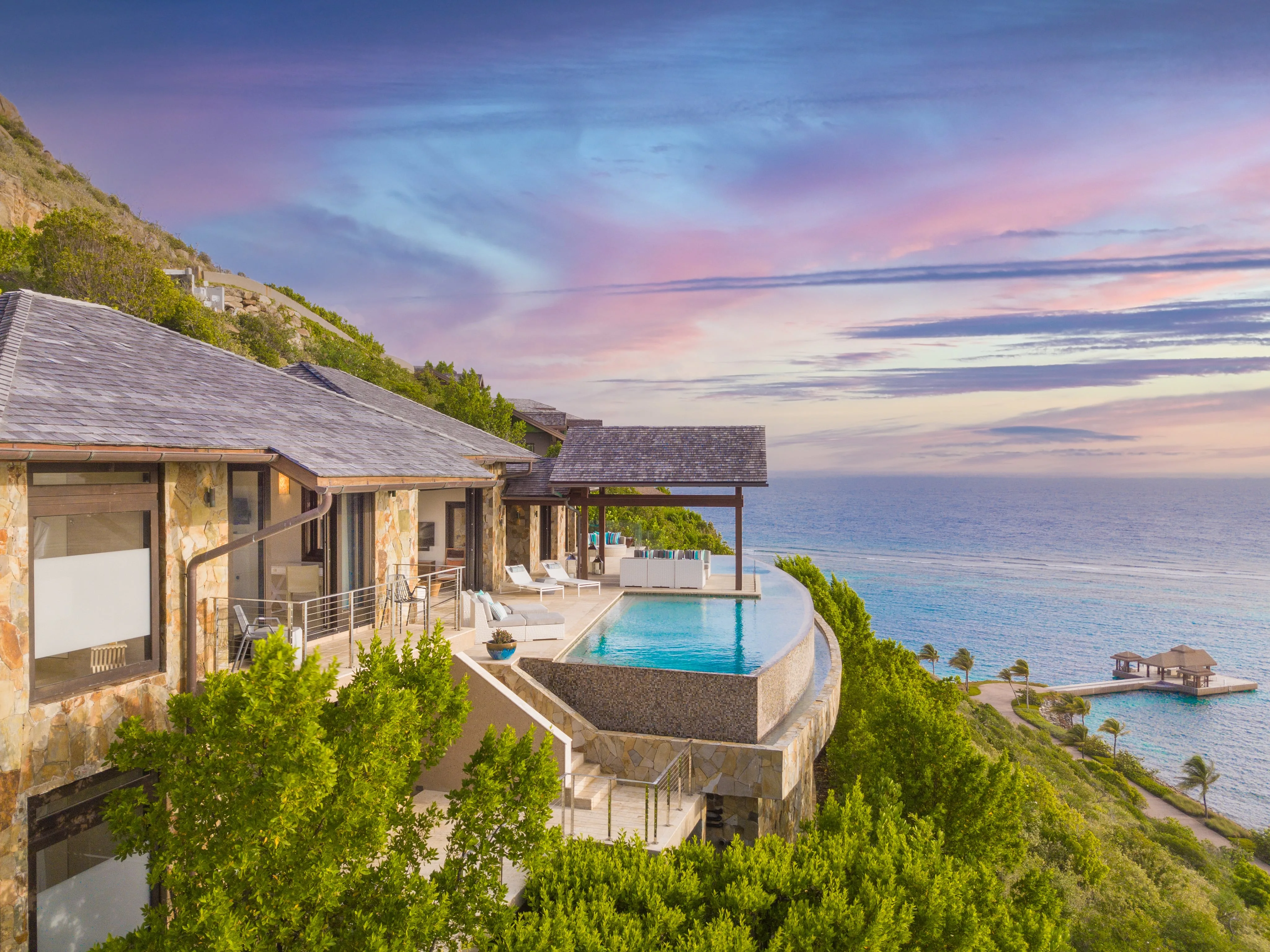 Luxurious seaside villa and coastal retreat with an infinity pool overlooking a serene ocean view. The sky is painted with pastel pink and purple hues at sunset, while lush greenery surrounds the stone-built villa, enhancing the tranquil atmosphere right at the water's edge.