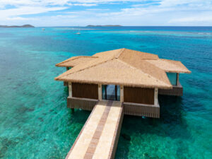 A thatched-roof overwater villa at Oil Nut Bay sits above clear turquoise ocean water in the British Virgin Islands, with boats and small islands visible in the distance under a partly cloudy sky.