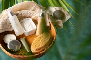 A wooden bowl holds VOYA skincare products, a body brush, a lit candle, and a textured cloth, set against lush green leaves for a natural, spa-like atmosphere reminiscent of the Oil Nut Bay spa.