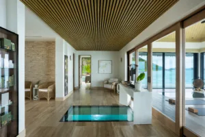 Modern, tropical villa interior with wood flooring, a central glass floor panel revealing water below—perfect for unwinding after a day at Oil Nut Bay spa. Large windows showcase ocean views, light furnishings, wooden ceiling, and lush greenery outside.