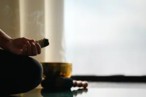 A person sitting cross-legged indoors holds a smoking bundle of sage in a tranquil scene reminiscent of an Oil Nut Bay spa. In the background, a singing bowl and mallet rest as soft natural light filters through sheer curtains.