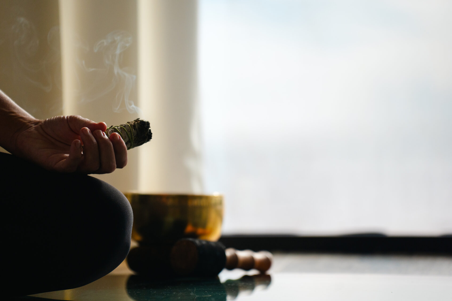 A person sits cross-legged indoors at Oil Nut Bay spa, holding a smoking bundle of sage. A singing bowl and mallet rest nearby, suggesting a tranquil scene of meditation or spiritual practice in soft, natural light.