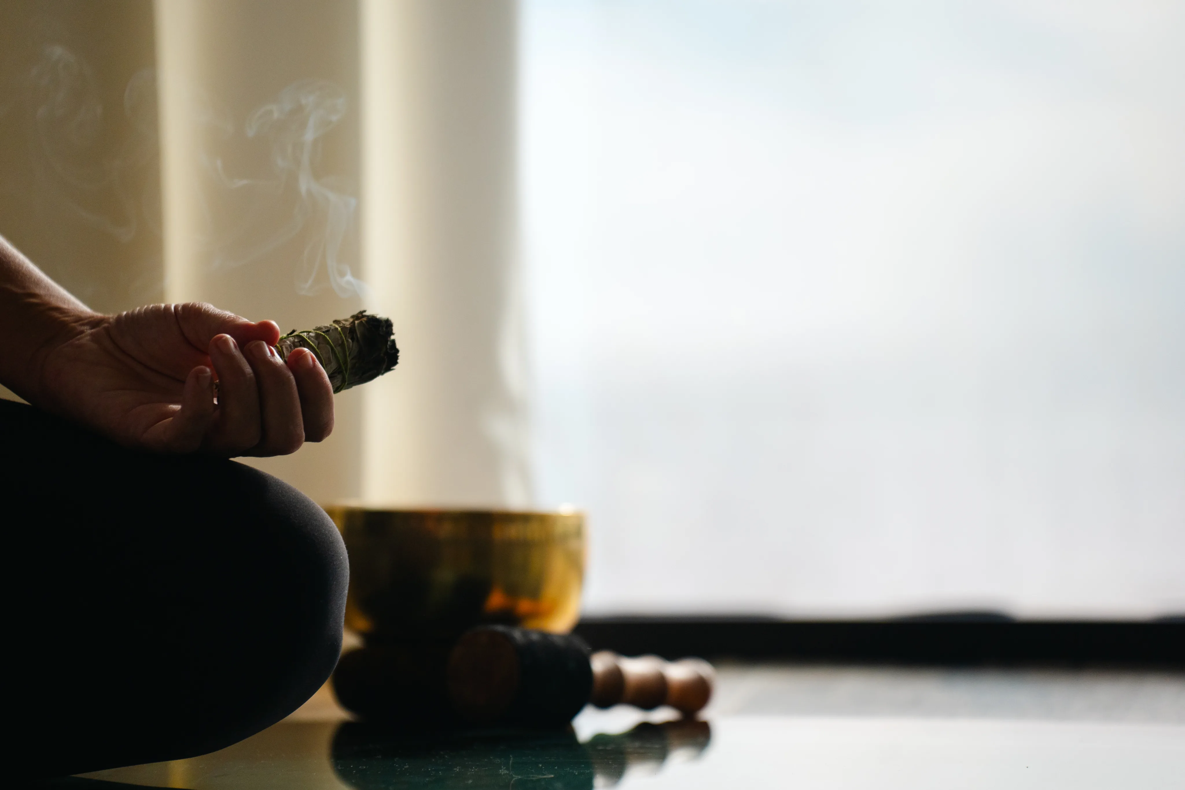 A person sits cross-legged indoors at Oil Nut Bay spa, holding a smoking bundle of sage. A singing bowl and mallet rest nearby, suggesting a tranquil scene of meditation or spiritual practice in soft, natural light.