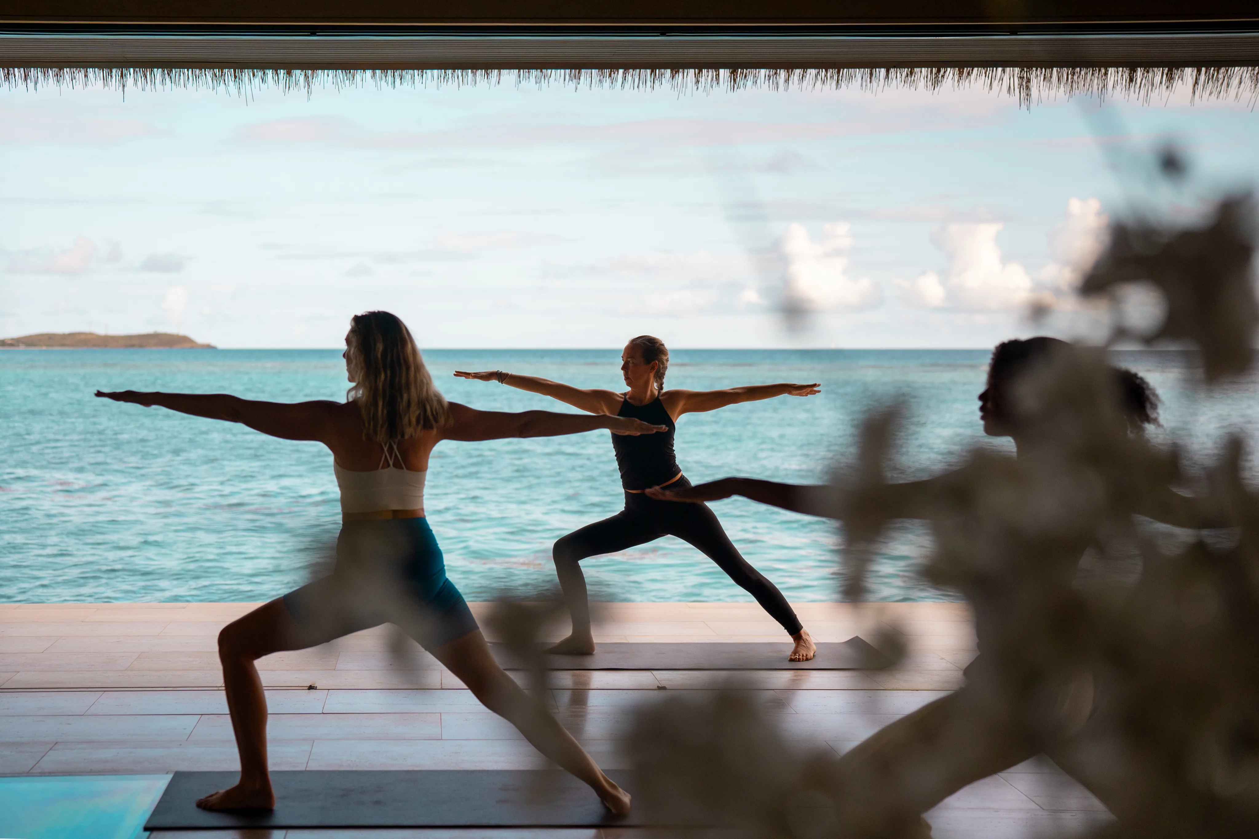 Three people practice yoga in a Warrior II pose on mats at Oil Nut Bay spa, in an open, covered space overlooking a calm, blue ocean under a partly cloudy sky.