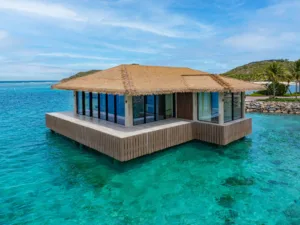 A modern overwater bungalow with a thatched roof sits above clear turquoise water at Oil Nut Bay in the British Virgin Islands, surrounded by lush greenery and blue sky.