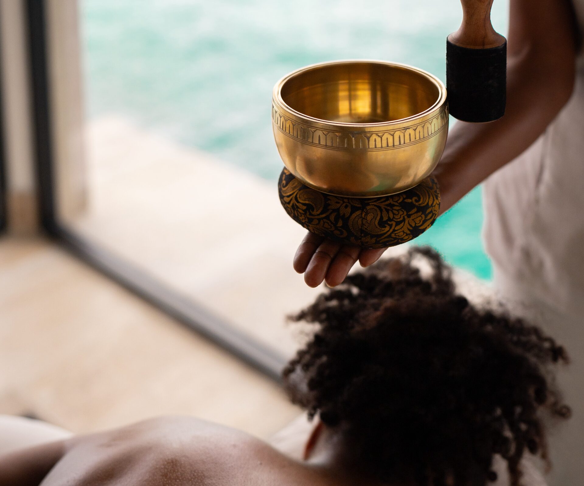 A person holds a singing bowl and mallet above someone lying down for a sound therapy session at Oil Nut Bay spa, with a blurred view of water and a window in the background.