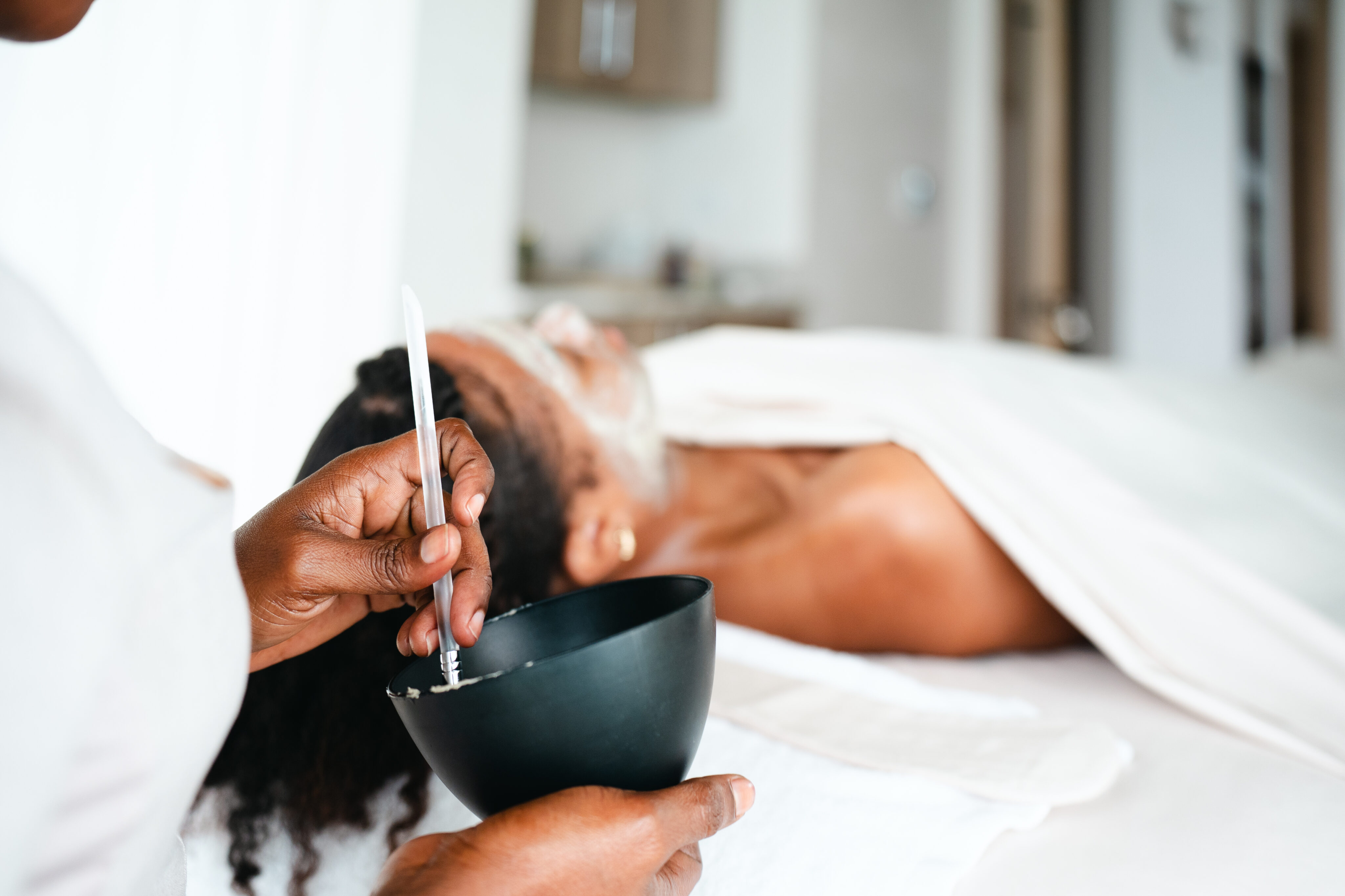 A person lies on a spa bed with a white towel, receiving a soothing facial at Oil Nut Bay spa. Another attendant mixes a facial mask in a black bowl, applying it gently in the calm and relaxing setting.