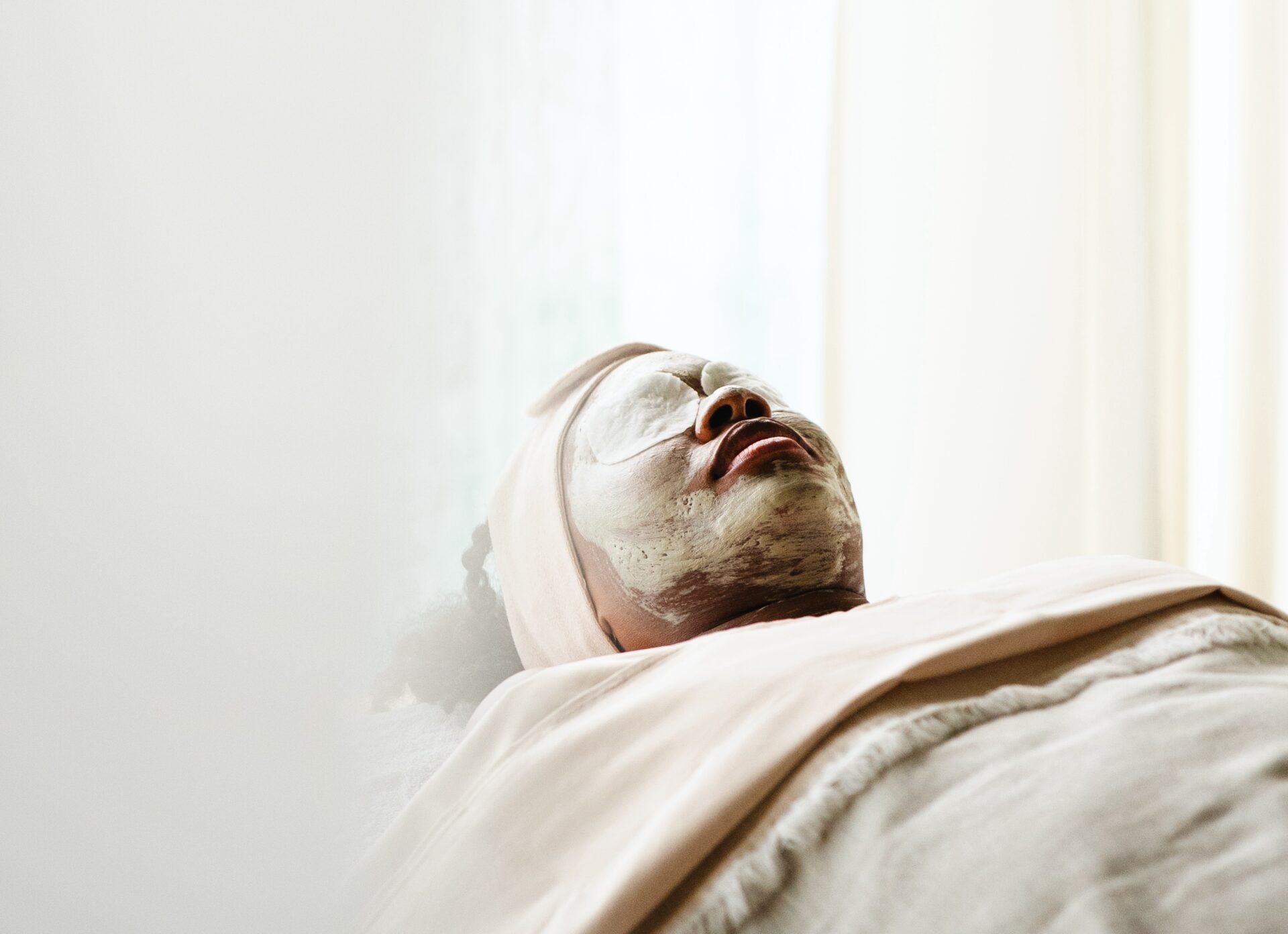 A person is lying down with eyes closed, wearing a headband and facial mask at Oil Nut Bay spa. They are covered with a beige blanket, relaxing in a softly lit, serene spa environment.