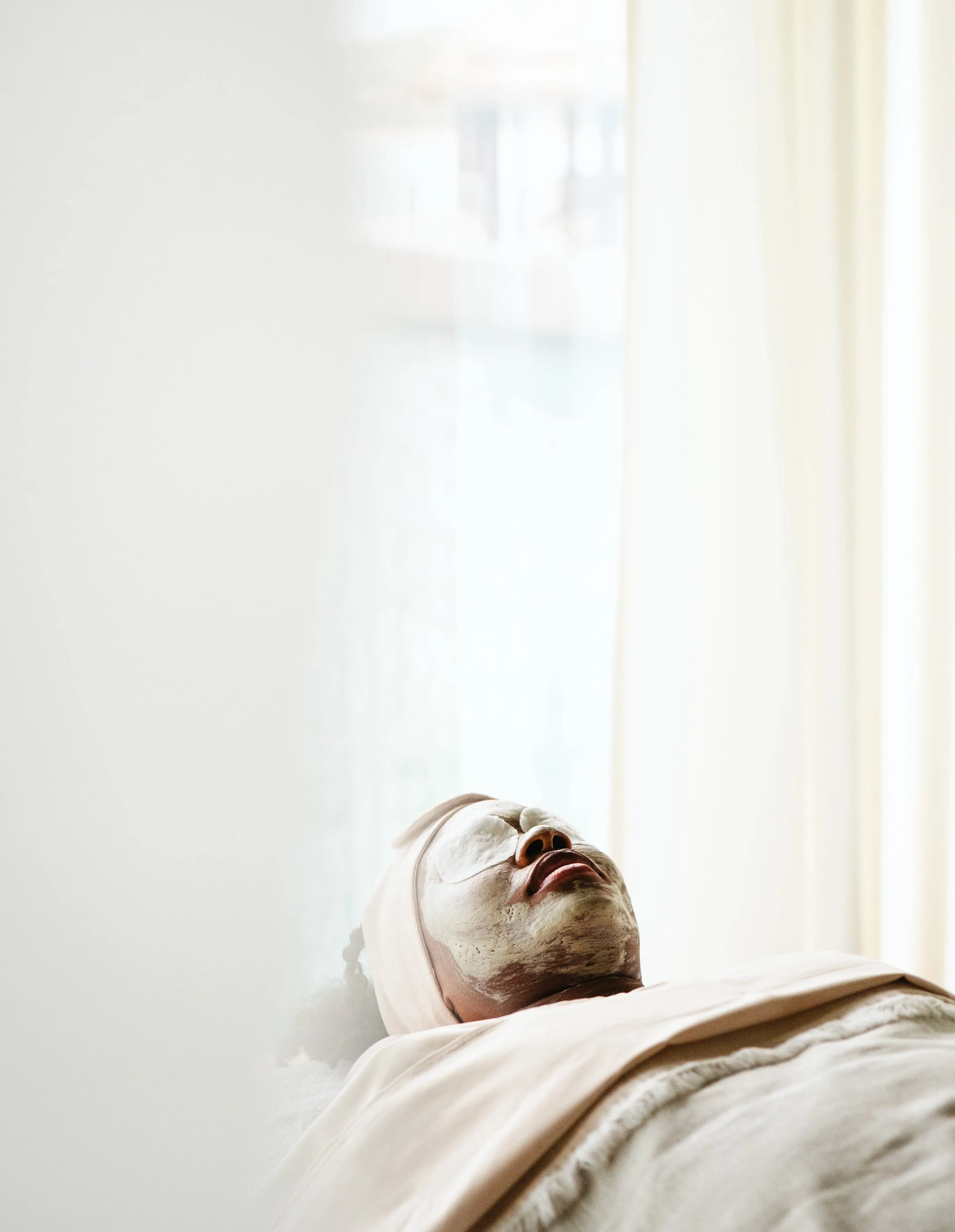 A person is lying down with a white facial mask applied, eyes closed, and covered with a light beige sheet in a softly lit, serene Oil Nut Bay spa setting.