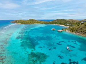 Aerial view of turquoise Oil Nut Bay in the British Virgin Islands, featuring coral reefs, a white sandy beach, green hills, floating docks, and a sailboat anchored in clear blue water under a partly cloudy sky.