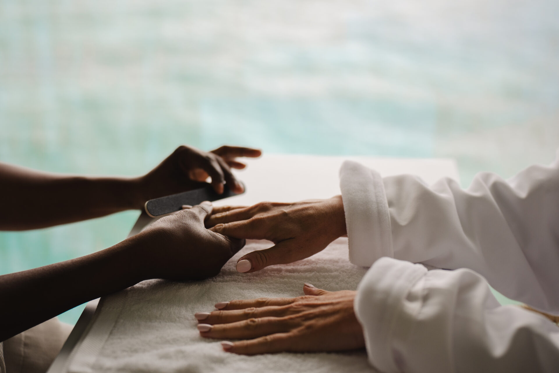 At Oil Nut Bay spa, a person in a white robe enjoys a manicure, their hands resting on a white towel as a nail technician expertly shapes their nails with a nail file.