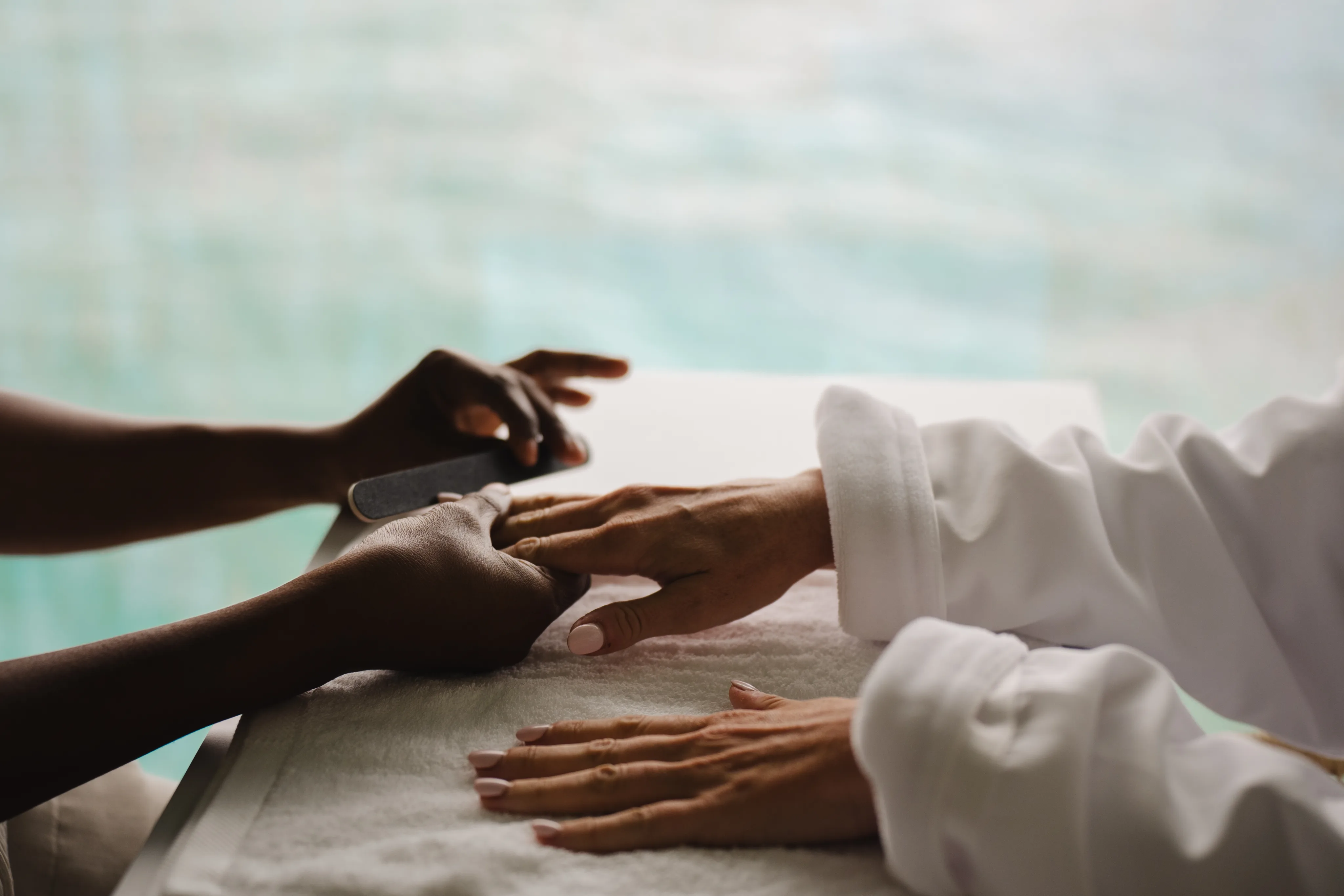 At Oil Nut Bay spa, a person in a white robe enjoys a manicure, their hands resting on a white towel as a nail technician expertly shapes their nails with a nail file.