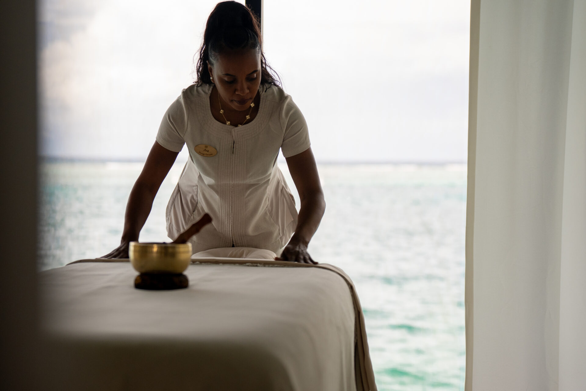 A spa therapist in a white uniform prepares a massage table in a serene room with large windows overlooking a calm ocean at Oil Nut Bay spa, with a singing bowl placed nearby.