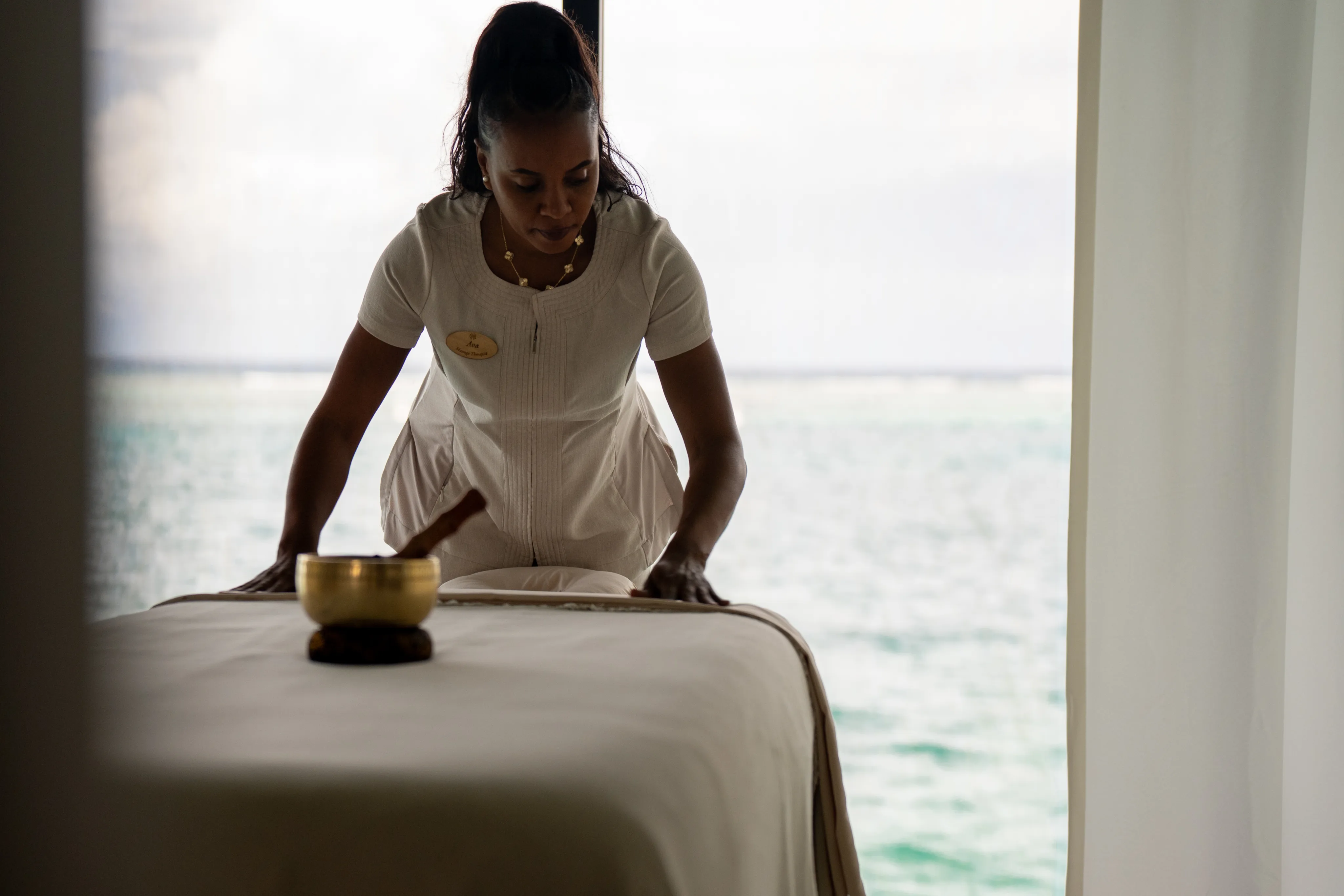 A spa therapist in a white uniform prepares a massage table in a serene room with large windows overlooking a calm ocean at Oil Nut Bay spa, with a singing bowl placed nearby.