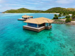 A thatched-roof overwater bungalow sits on clear turquoise water at Oil Nut Bay, surrounded by lush green hills and palm trees on a tropical island in the British Virgin Islands. Another bungalow is visible in the distance.